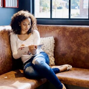Woman sitting on a café bench by a window writing in a notebook in natural light.