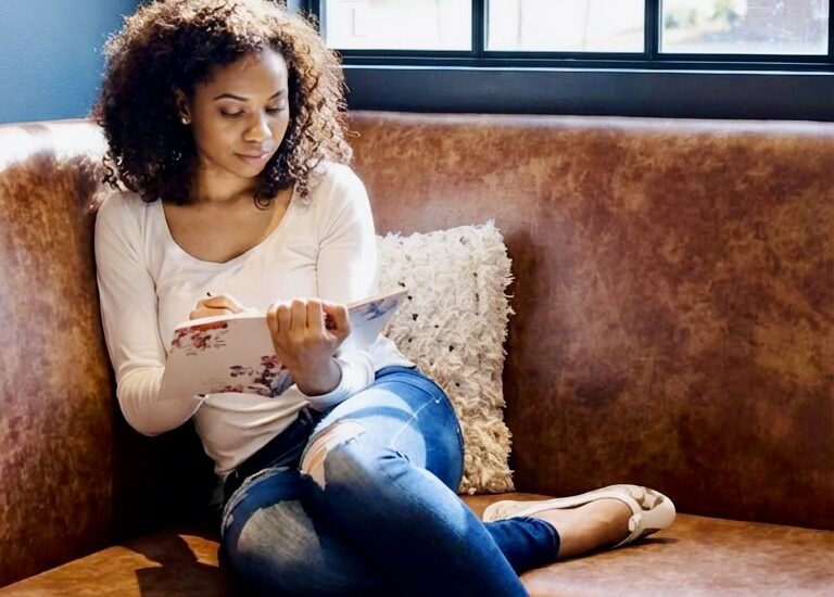 Woman sitting on a café bench by a window writing in a notebook in natural light.